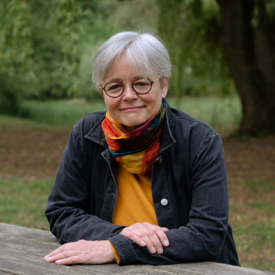 Dr. Annegret Haage lehnt draußen an einen Holz-Tisch, Bäume im Hintergrund 